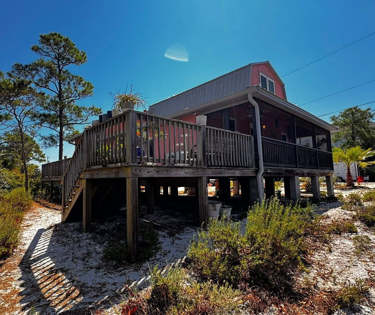 A pink elevated house with a wooden deck, set on sandy ground among trees under a bright blue sky.