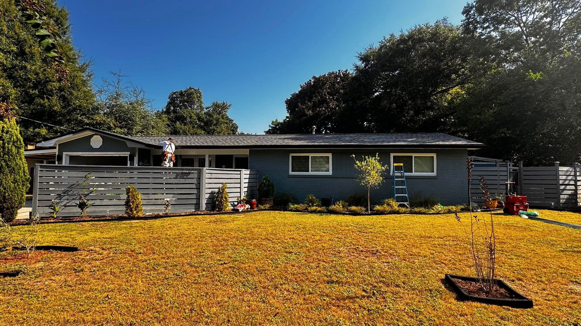A one-story gray house with a horizontal fence and a person on a ladder working on the roof, viewed across a grassy lawn.