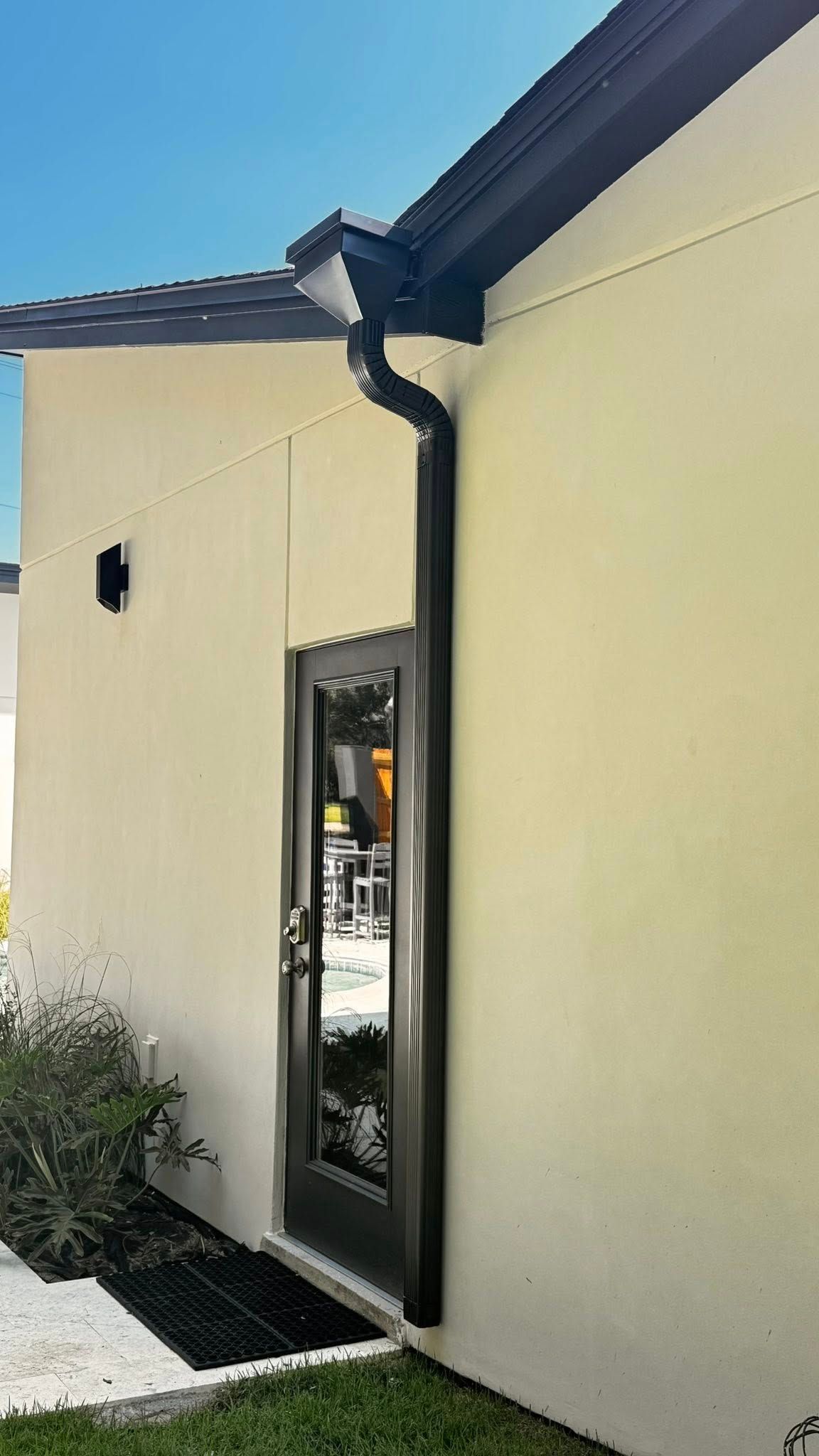 A dark downspout runs vertically along the wall, obstructing the edge of a glass door on a light-colored building.