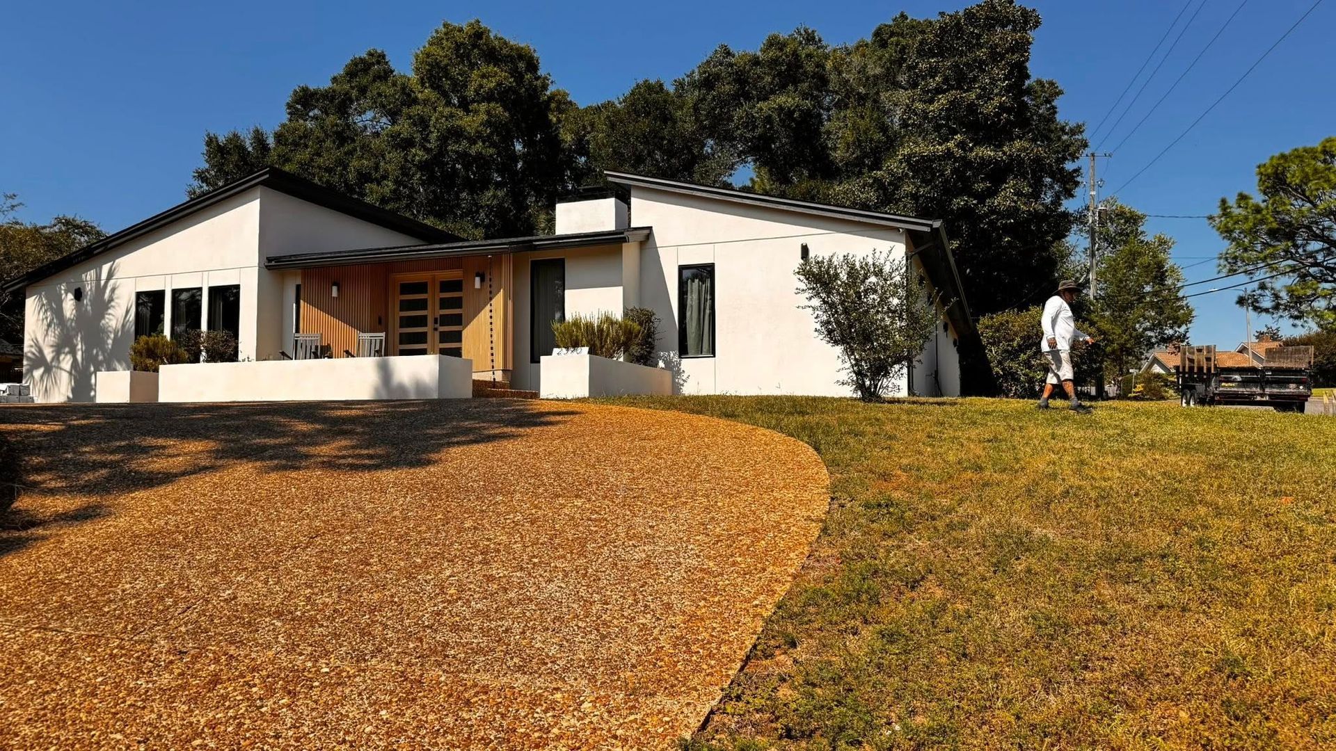 A white, single-story house with a gravel driveway and surrounding trees under a clear blue sky.