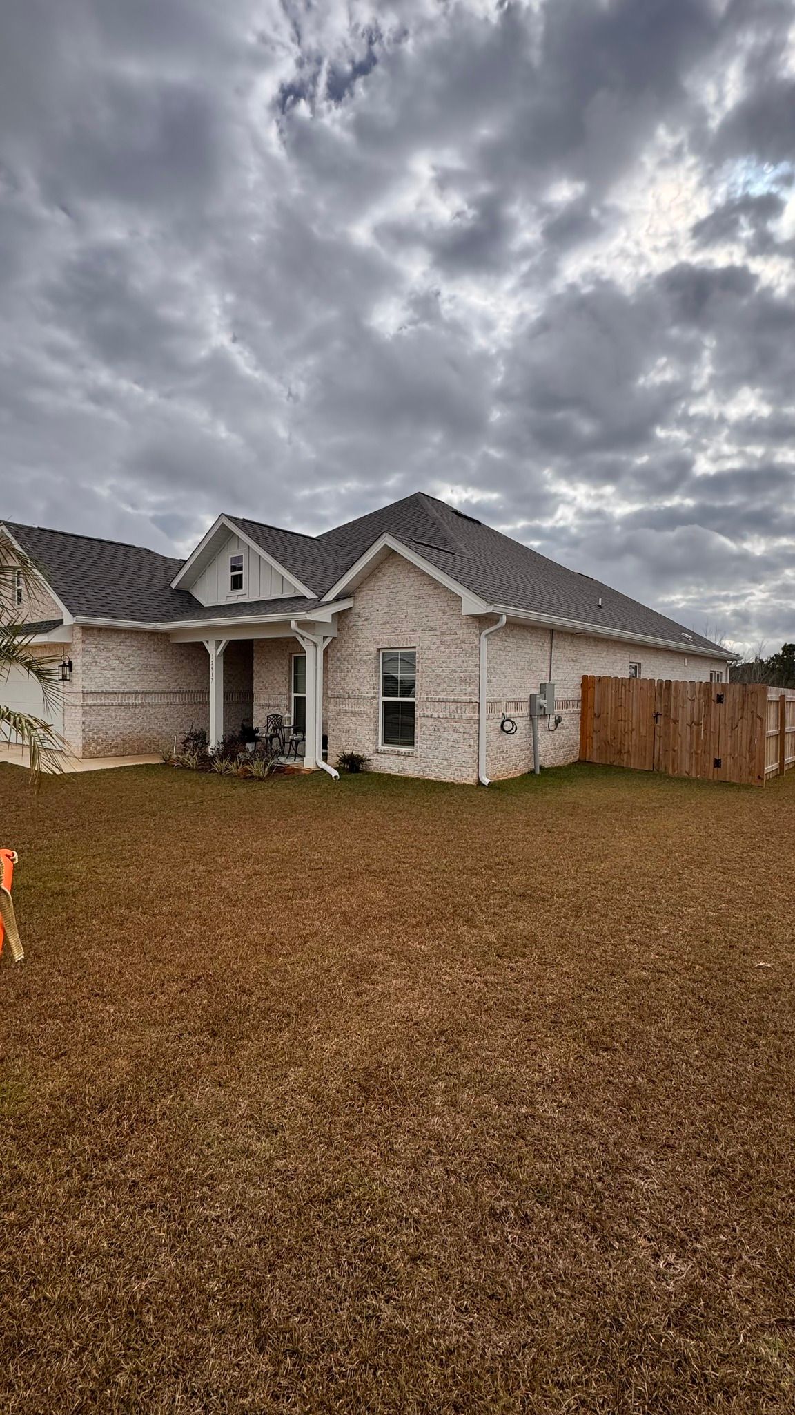 A single-story, light-colored brick house with a dark shingled roof under a cloudy sky, with a wooden fence on the right.