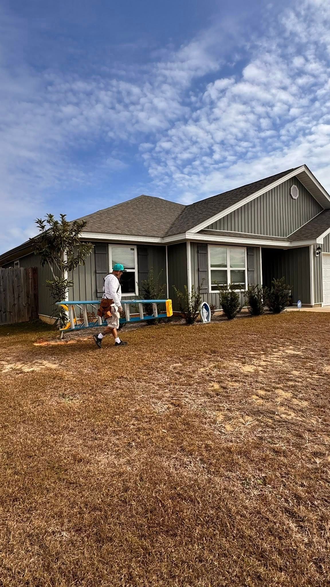 A person carrying a multi-purpose ladder across the lawn in front of a gray single-story house under a cloudy blue sky.