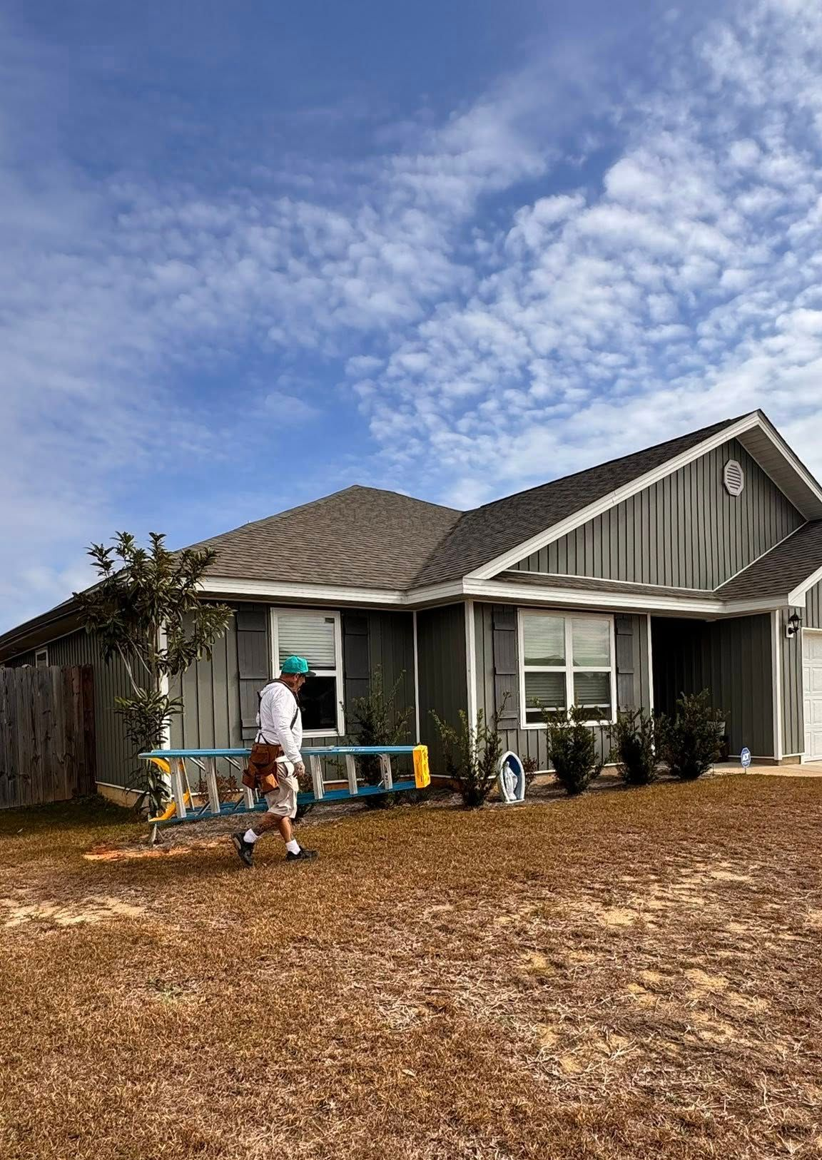 A person carrying a blue ladder across the front yard of a single-story gray house under a cloudy blue sky.