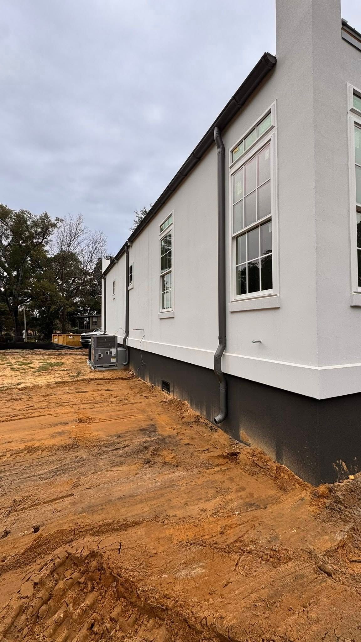 A side view of a white building with dark trim and foundation, set against a dirt lot under an overcast sky.