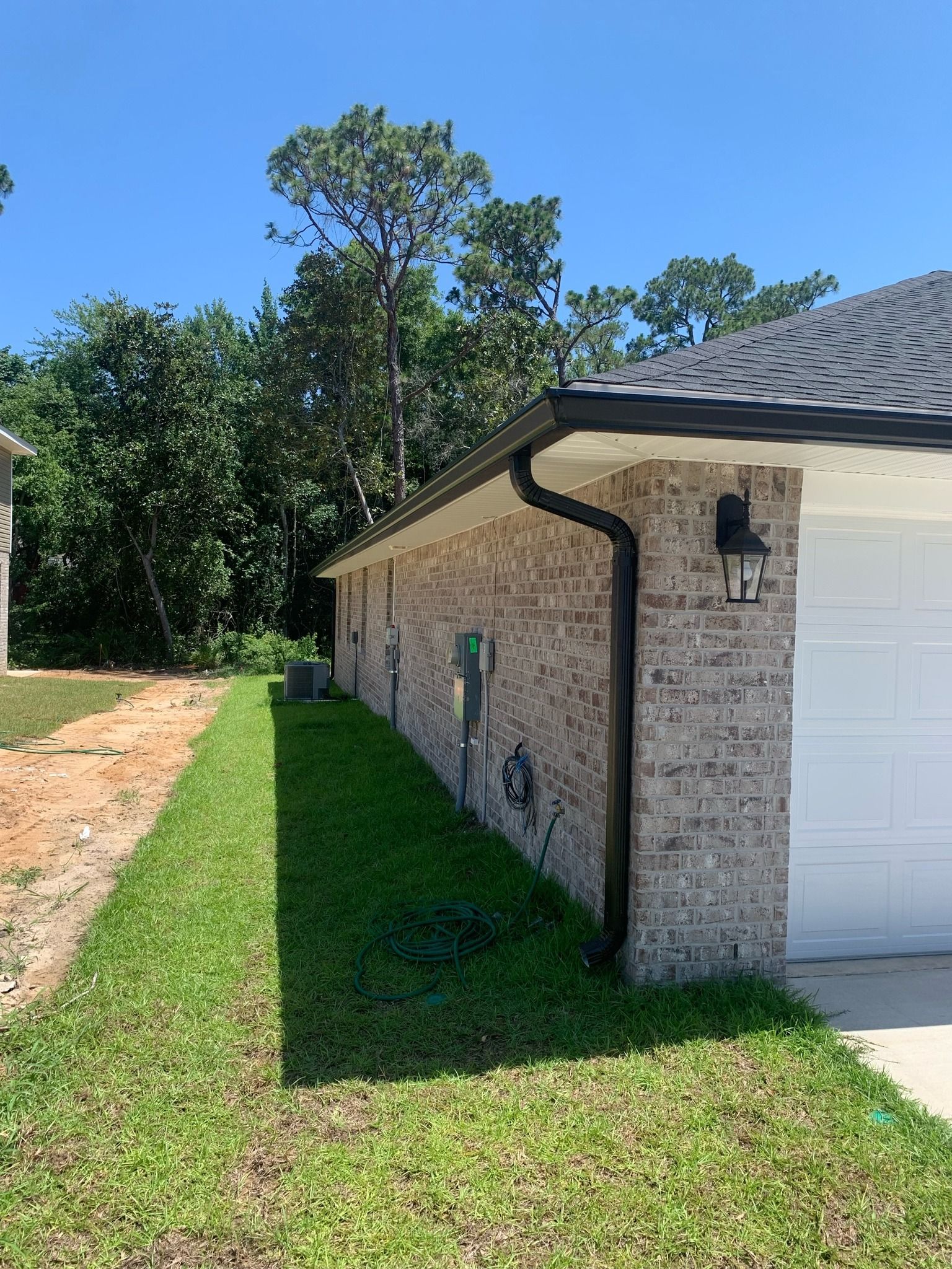 A view of a tan brick house exterior with black gutters and a garage, set against a backdrop of green trees.