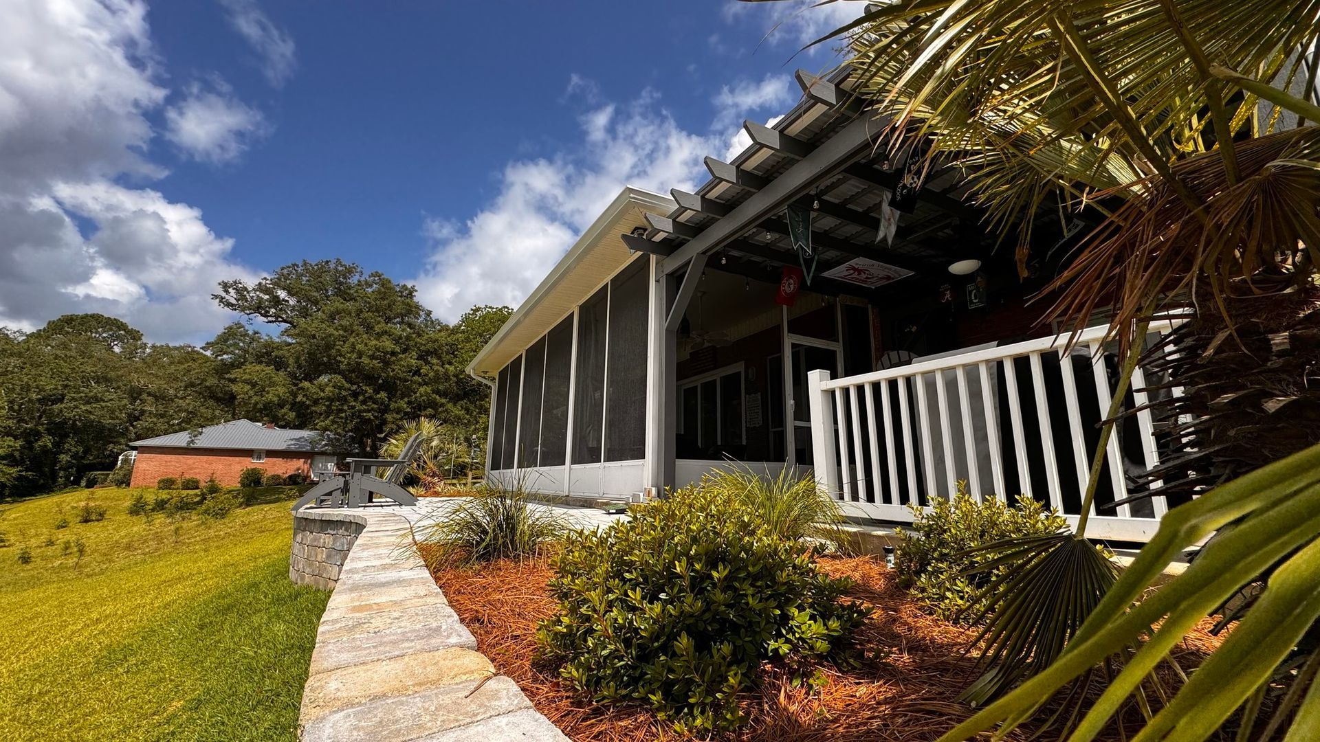 A sunlit patio with a screen enclosure and white railing, featuring a stone retaining wall and palm fronds under blue skies.