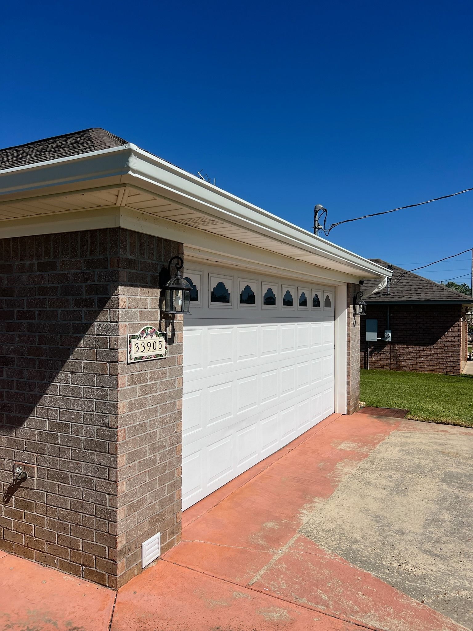 A corner view of a brick house garage with a white door, decorative windows, a wall lantern, and a concrete driveway.