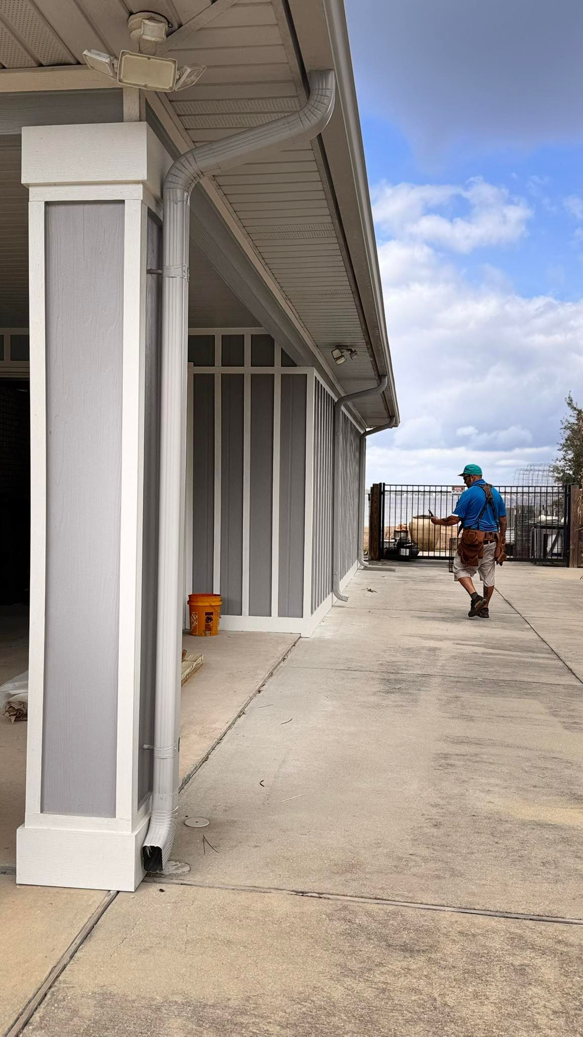 A worker in a blue shirt walks along a concrete walkway beside a building exterior with white pillars and gray siding.