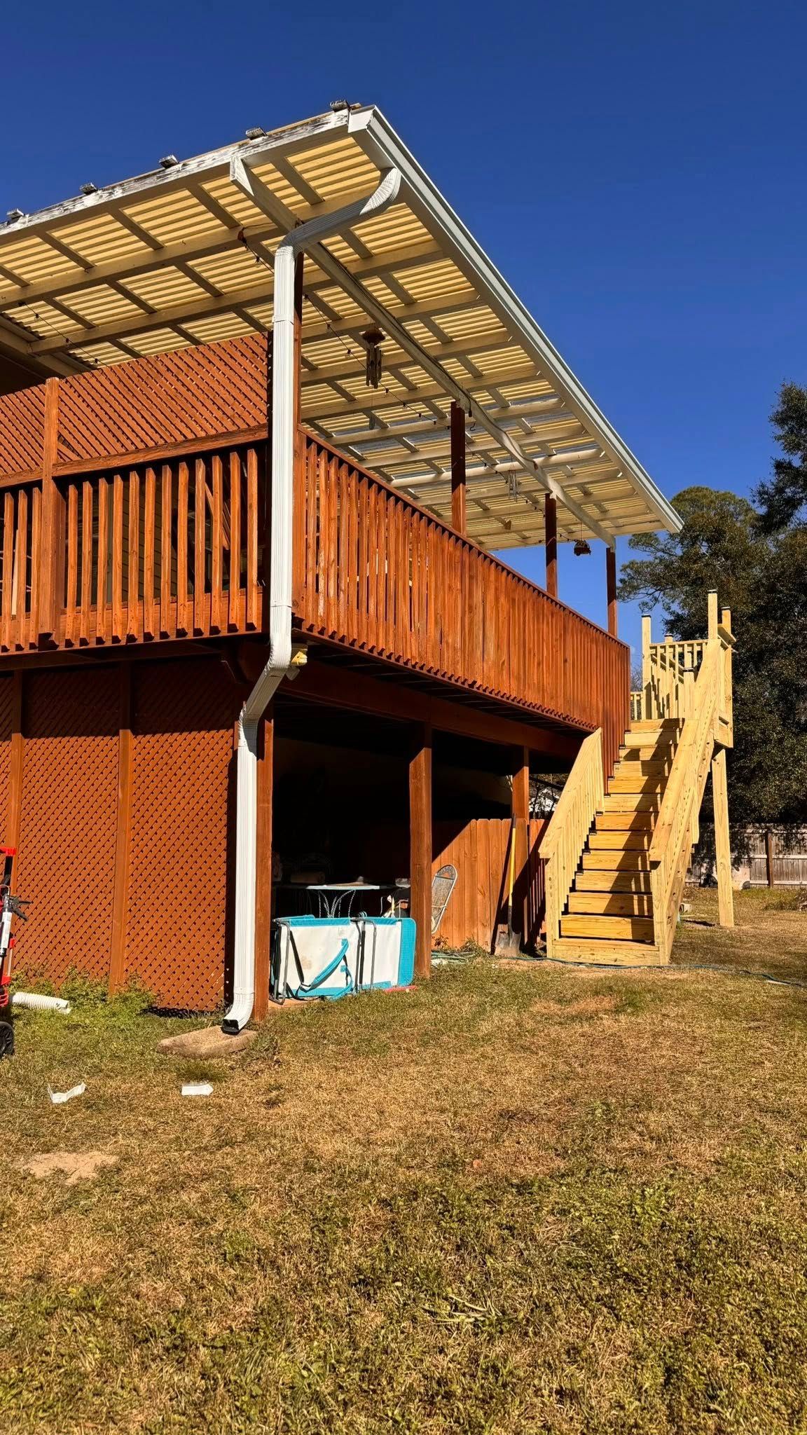 A raised wooden deck with a white slatted patio cover, lattice skirting, and wooden stairs leading up, set in a grassy yard.