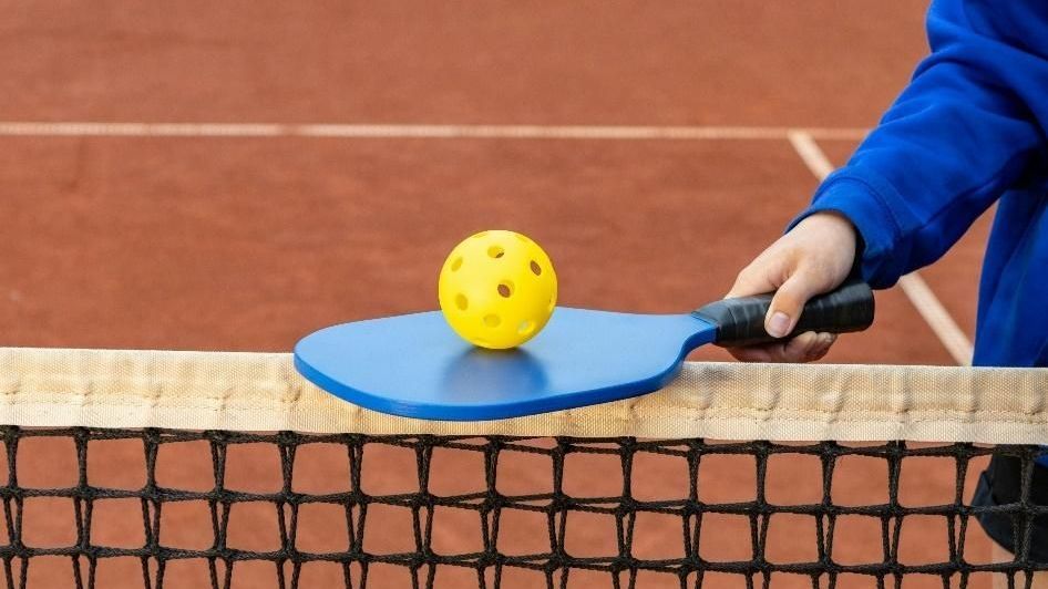 A person is holding a paddle with a yellow ball on it over a tennis net.