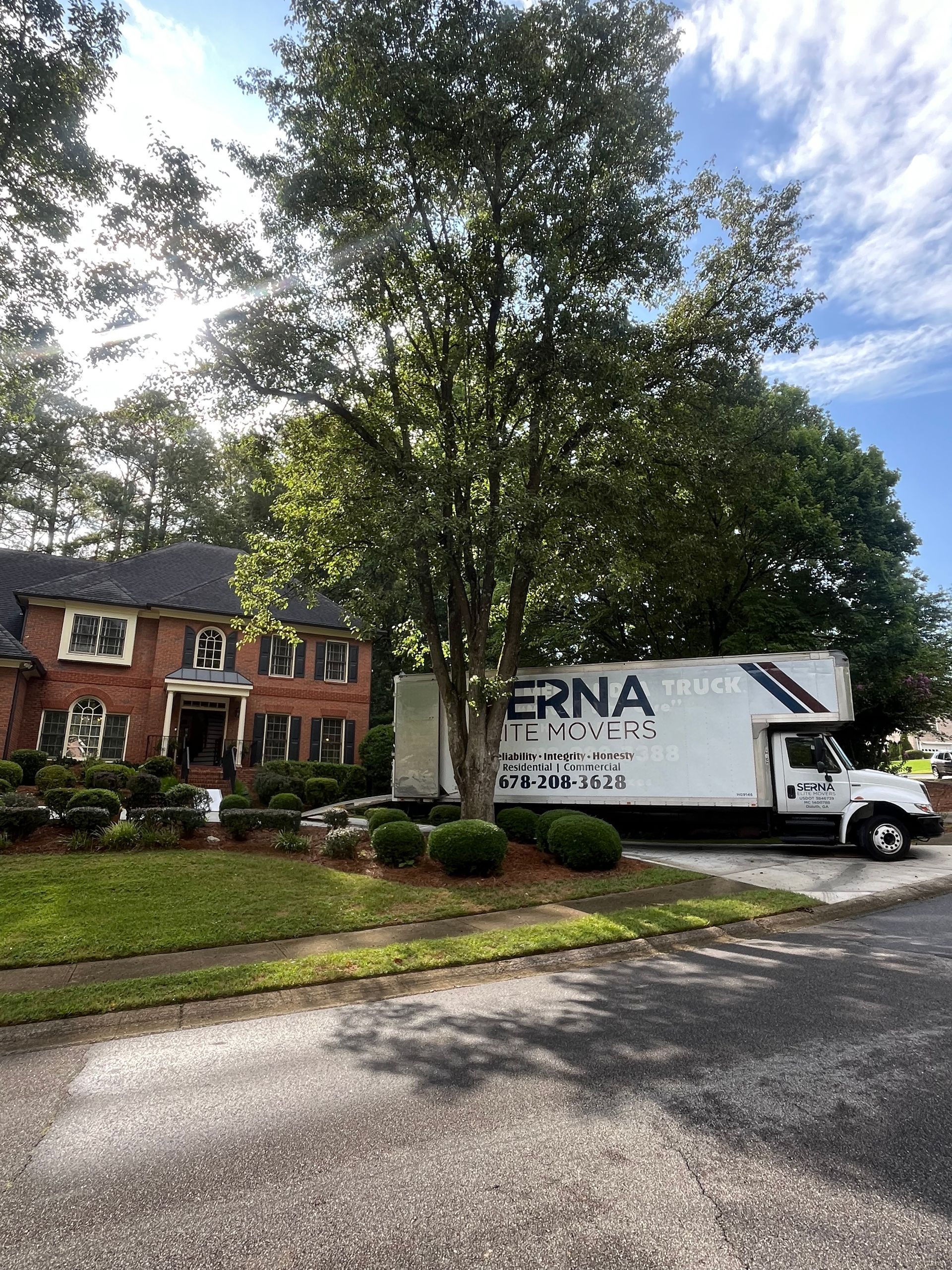 Moving truck parked in front of a brick house, green trees, blue sky.