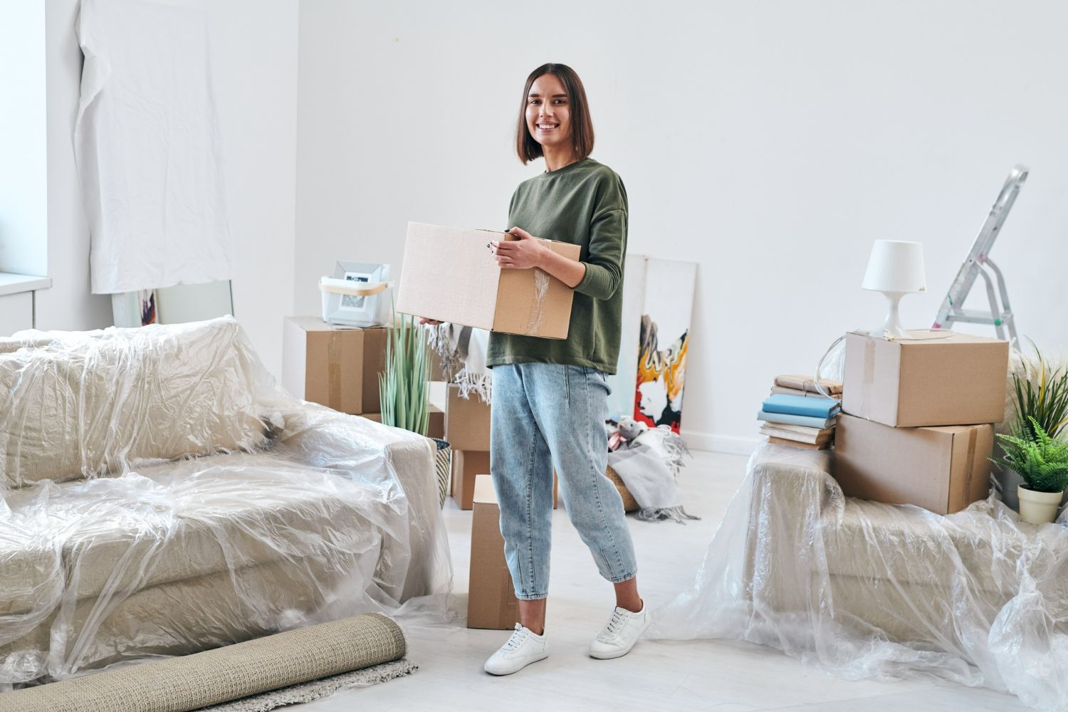 Person holding a box in a bright, unpacked living room with moving boxes and covered furniture