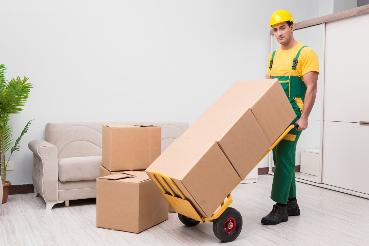 Moving worker using a hand truck to move boxes in a room, wearing green overalls, yellow shirt and hard hat.