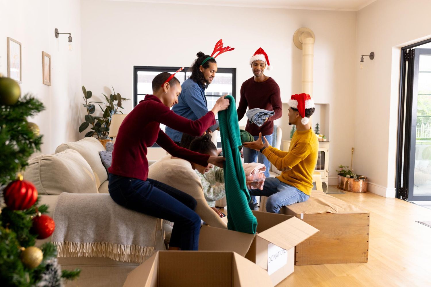 People in a living room unpacking Christmas decorations; one wearing reindeer antlers, others wearing Santa hats.