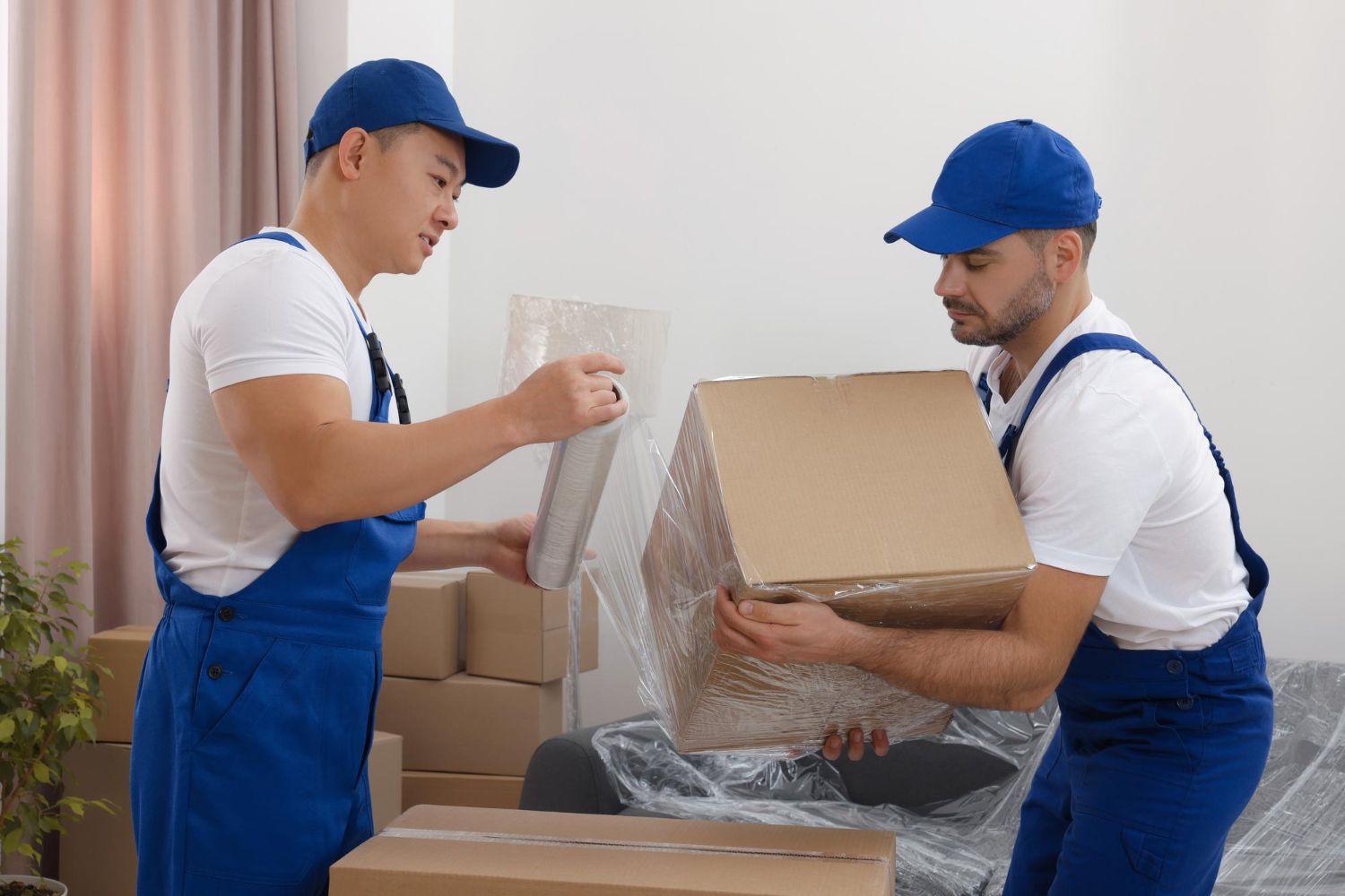 Two movers wearing blue uniforms pack a box, preparing for a move in a bright room.