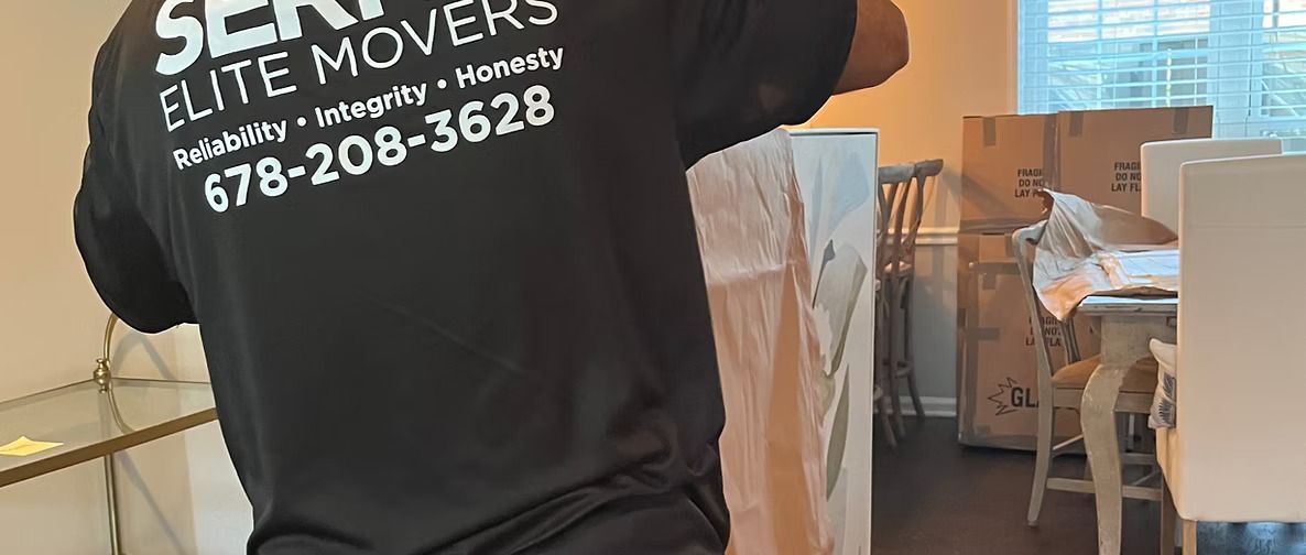 A person wearing a SERVE ELITE MOVERS t-shirt packs furniture in a house with boxes and a table.