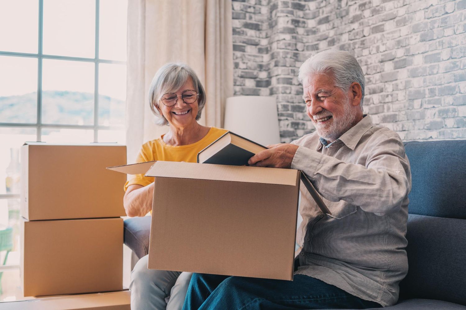 Smiling older couple unpacking books from a cardboard box on a couch, indoors.