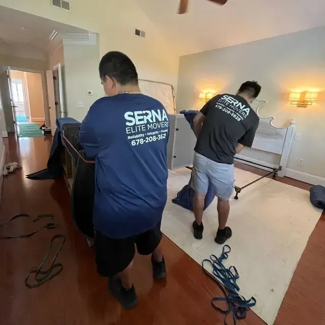 Two movers in blue shirts carry furniture through a living room with tools and cords on the floor.