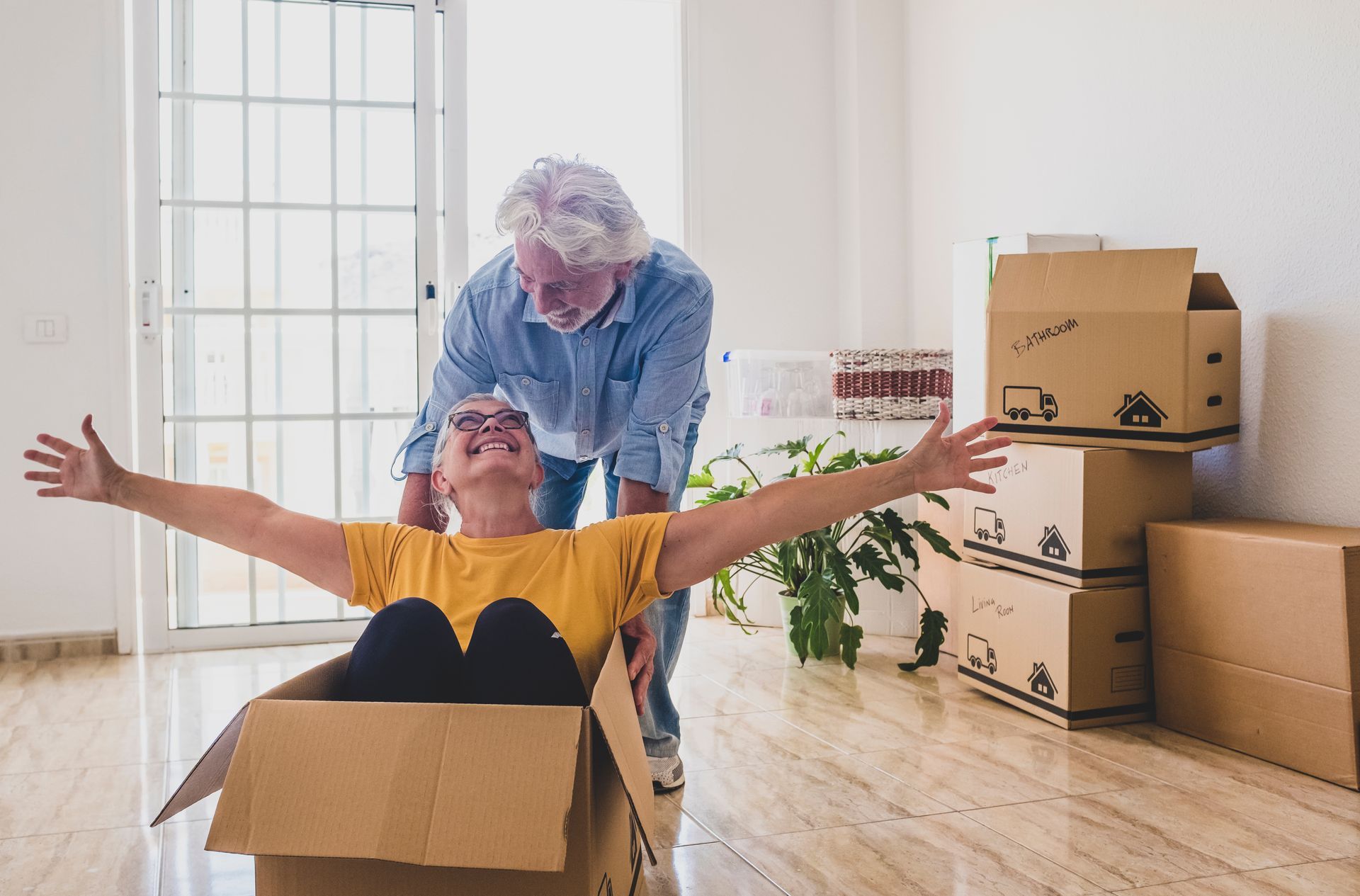 Elderly couple in new home: woman in box with arms out, man pushing, smiling; moving boxes in background.