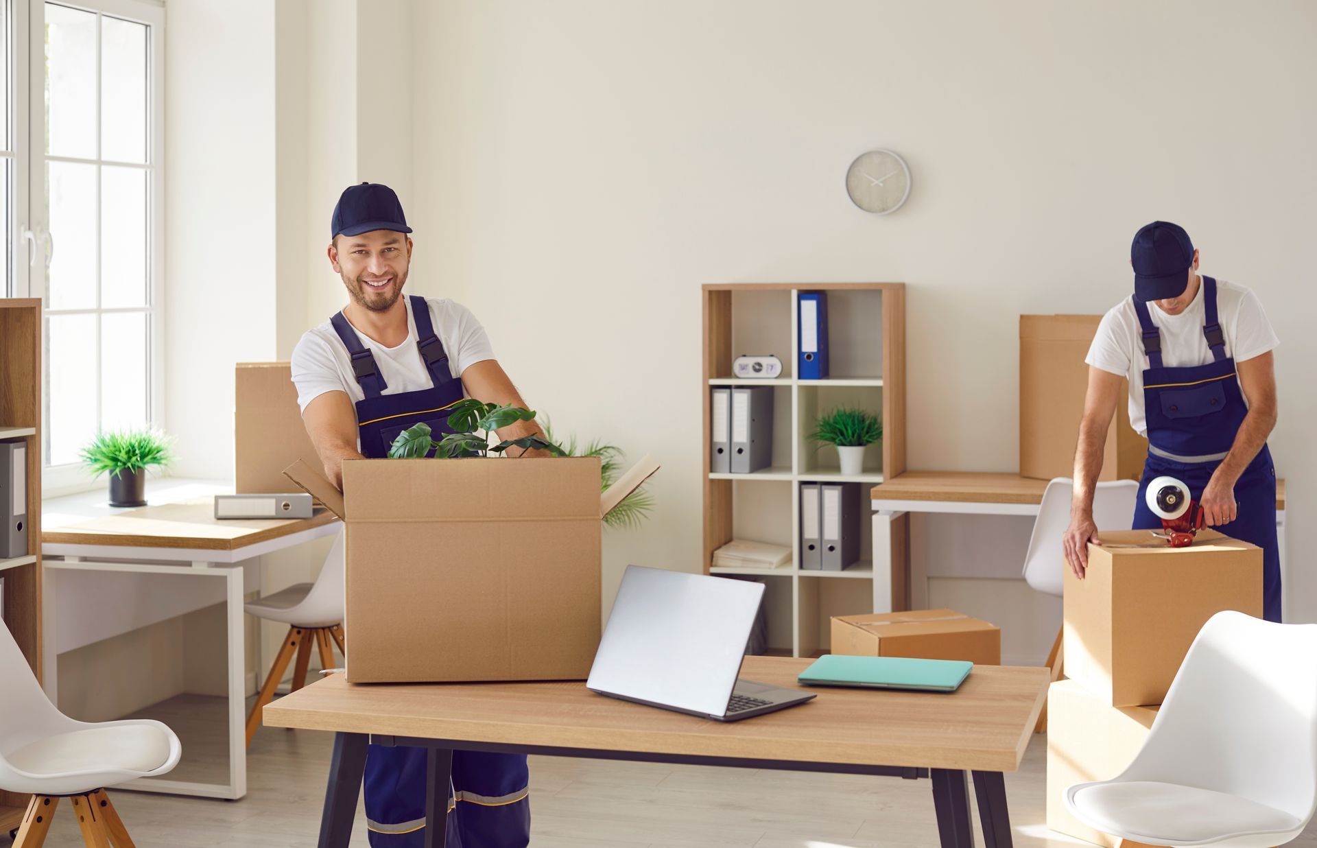 Movers packing office. One smiling, holding box. Other sealing box. Desks, shelves, white walls, natural light.