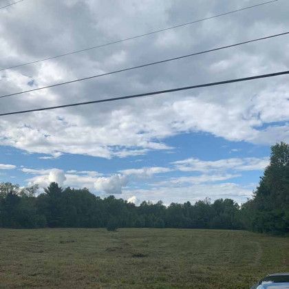 A car is parked in a field with trees in the background and a blue sky with clouds.