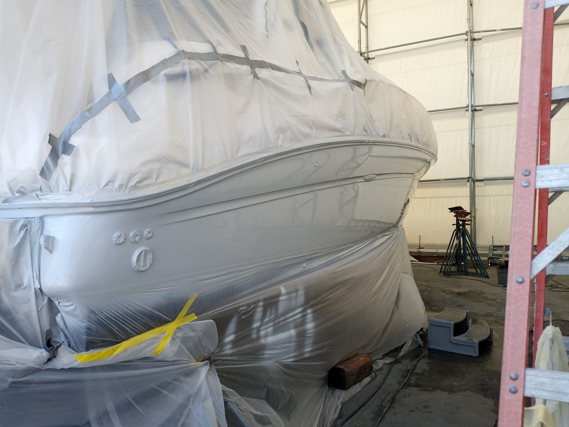 Boat covered in plastic sheeting, likely for painting, in a boatyard or workshop.