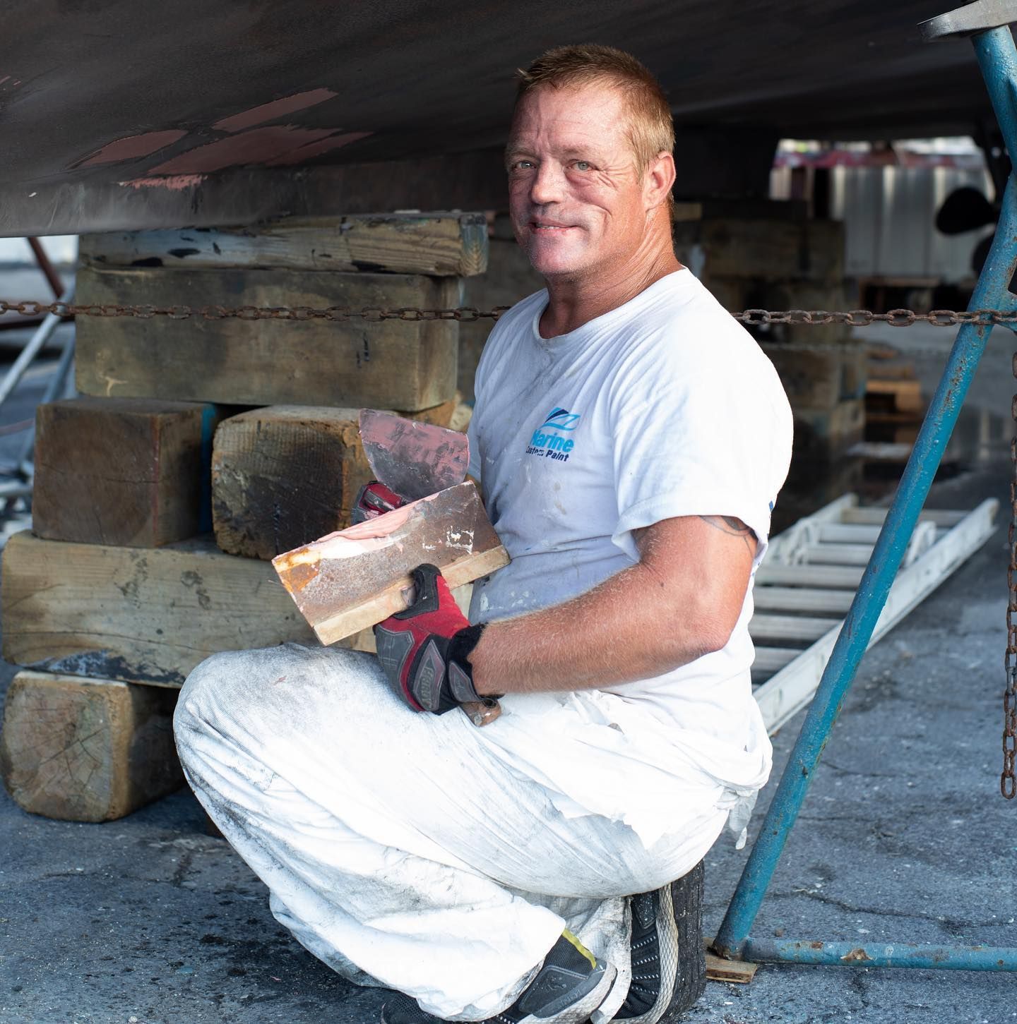 Man in white overalls squats, holding wood, near a boat in a shipyard.