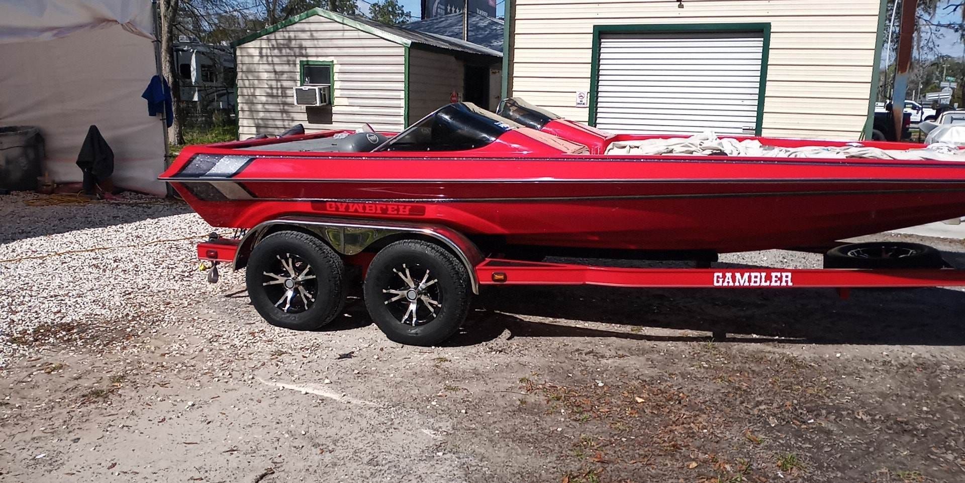 Red boat on a trailer, with black wheels, parked on gravel, near buildings.