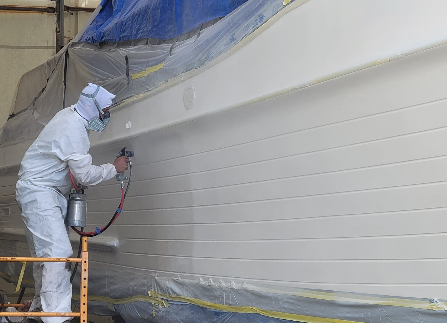Person in protective suit spray-painting a white boat in a workshop.