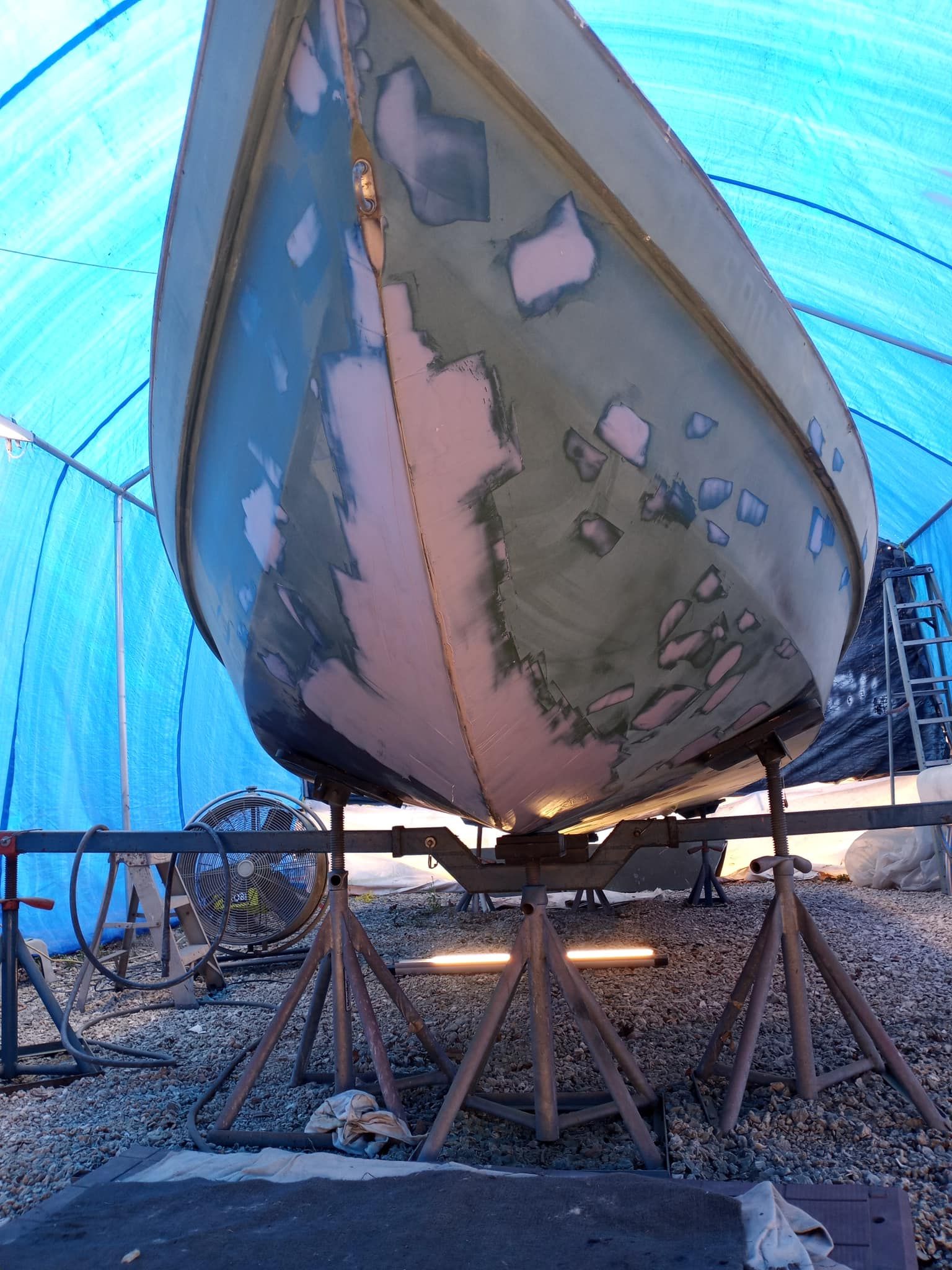 Boat hull on stands, undergoing repair with patches of pink and green primer, under a blue tarp.