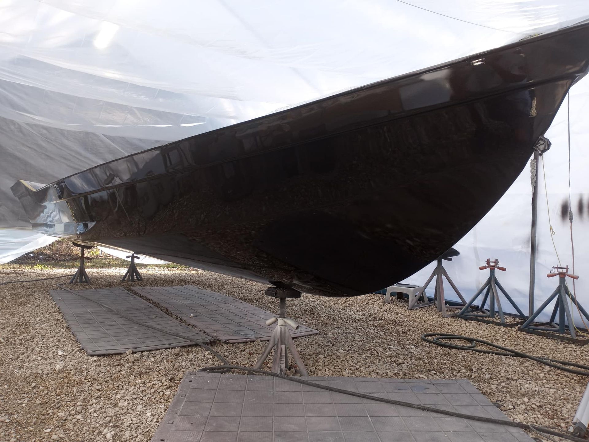 Black boat on stands, under a transparent cover in a gravel lot.