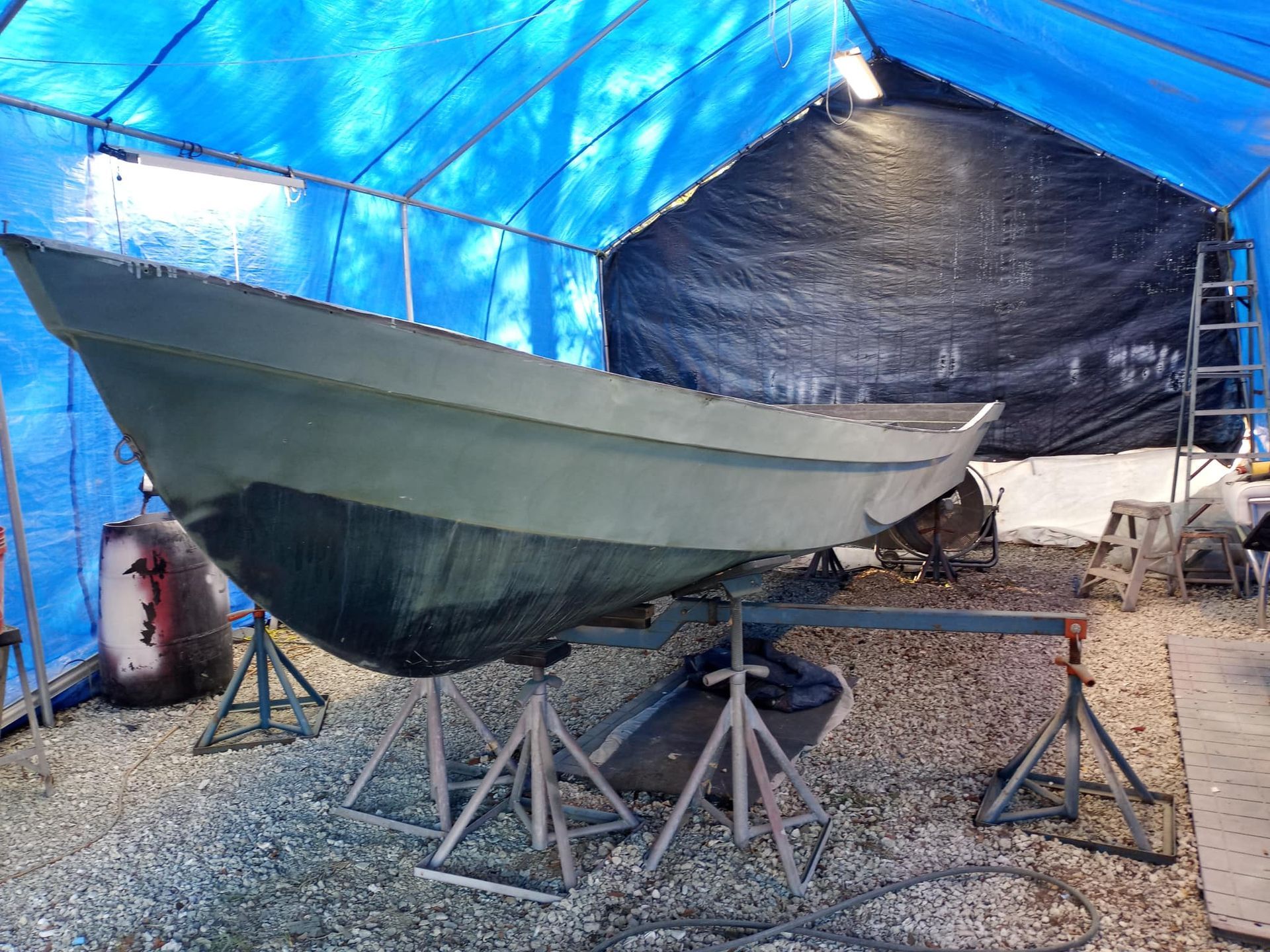 Boat on stands inside a blue tarp shelter; undergoing repairs, grey hull, metal supports.