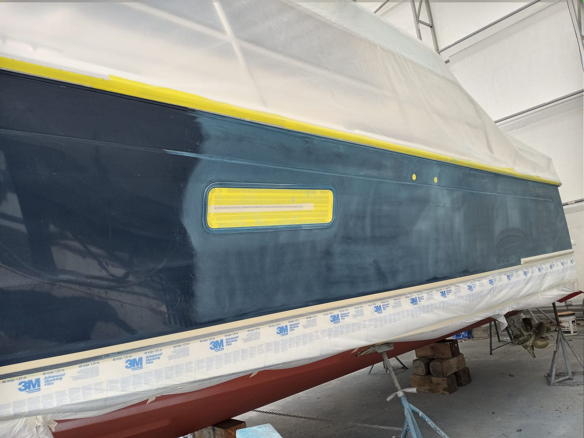Boat hull being painted; blue hull with masking tape and protective covering in a workshop setting.