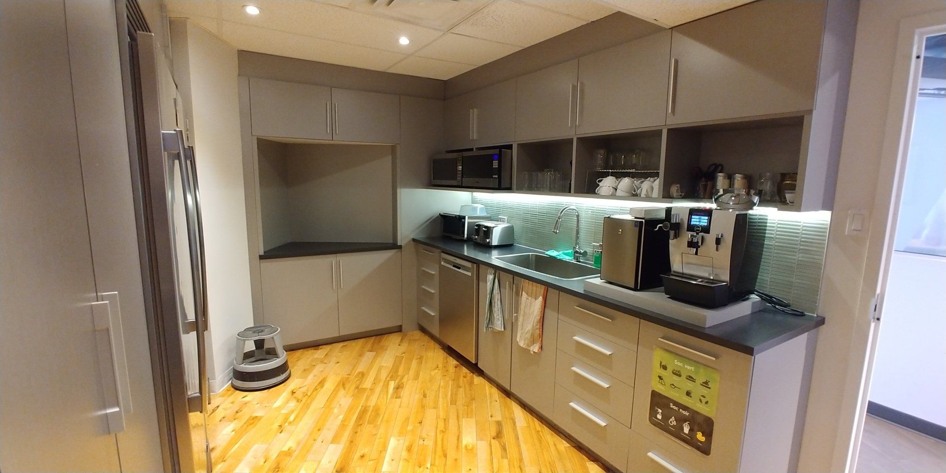 Une cuisine avec des appareils électroménagers en acier inoxydable, des armoires grises et du parquet. Une machine à café et un micro-ondes sont visibles.