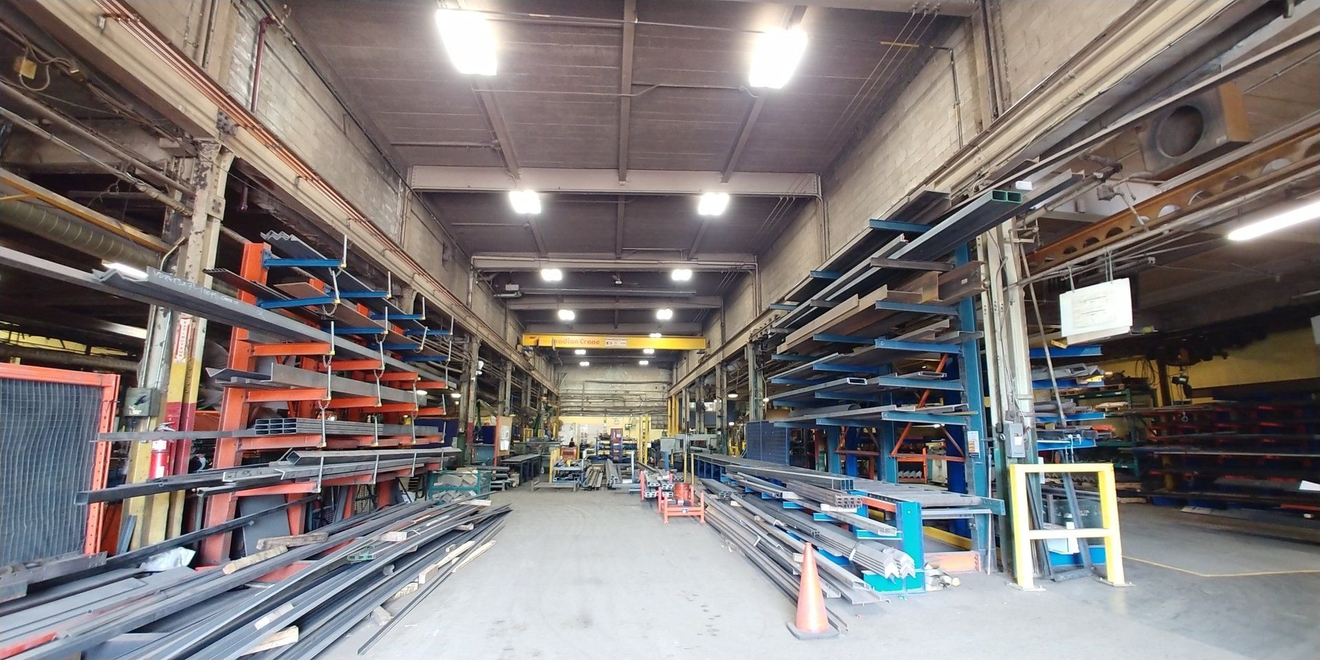 Intérieur d'entrepôt rempli de matériaux métalliques sur des étagères ; un pont roulant est visible au loin.