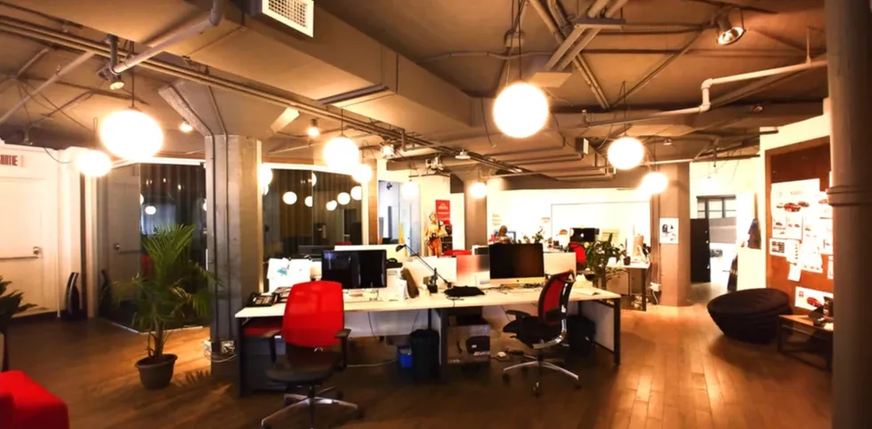 Un bureau moderne à aire ouverte avec des bureaux, des chaises rouges, des suspensions lumineuses en forme de globe et un plafond industriel avec des tuyauteries apparentes.