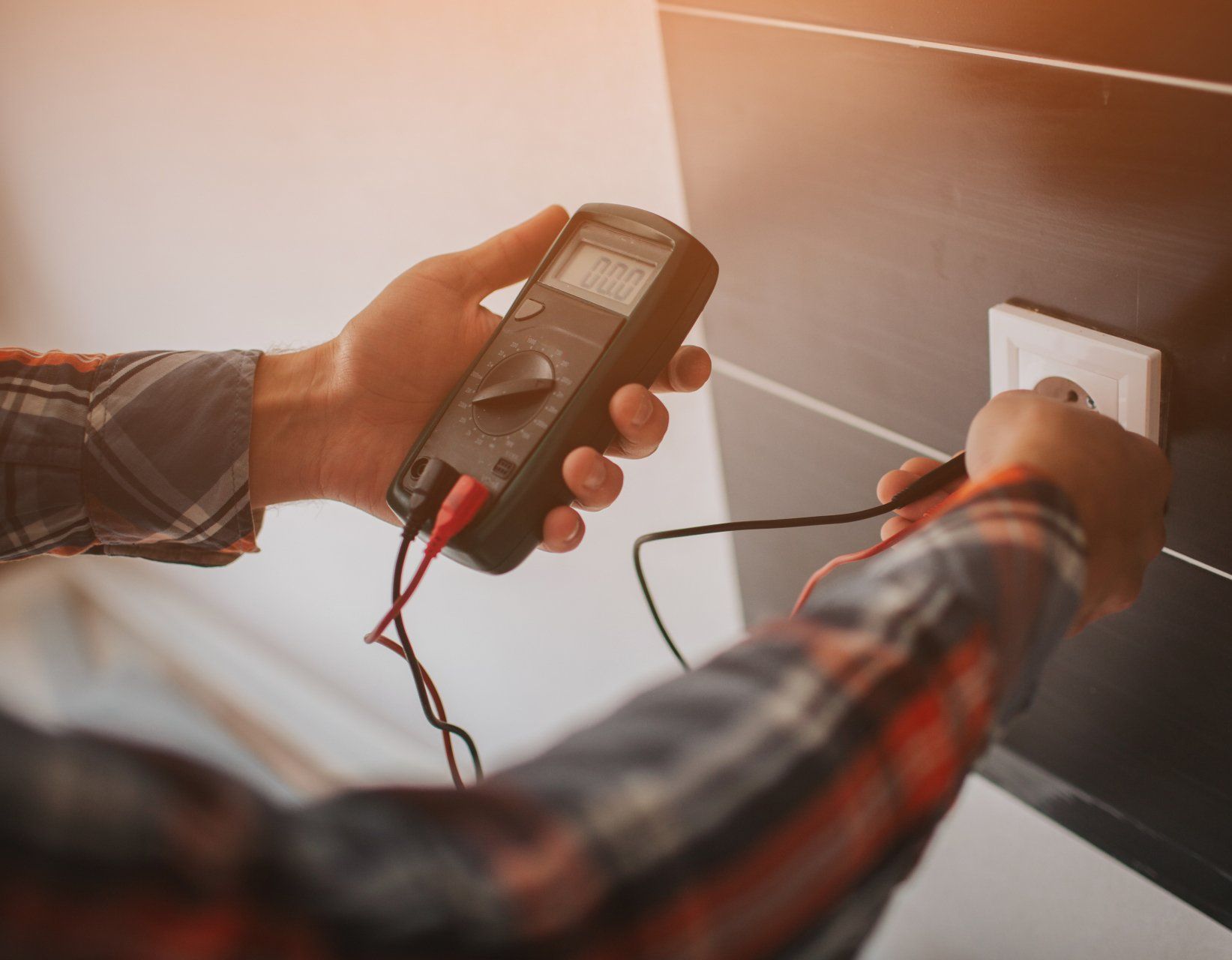 Des mains utilisent un multimètre pour tester une prise de courant sur un mur sombre ; l'électricien porte une chemise à carreaux.