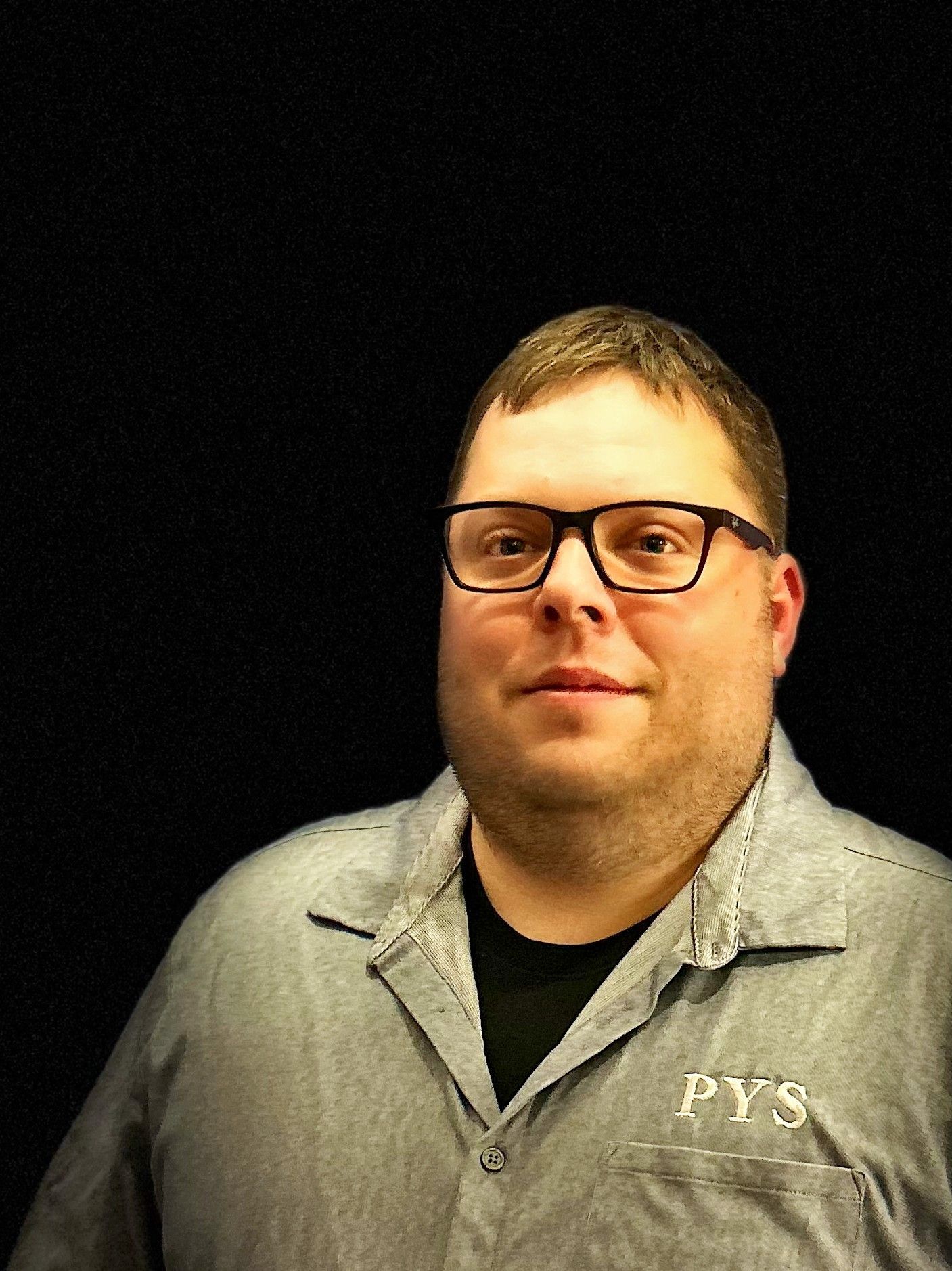 A man wearing glasses and a grey shirt is standing in front of a black background.