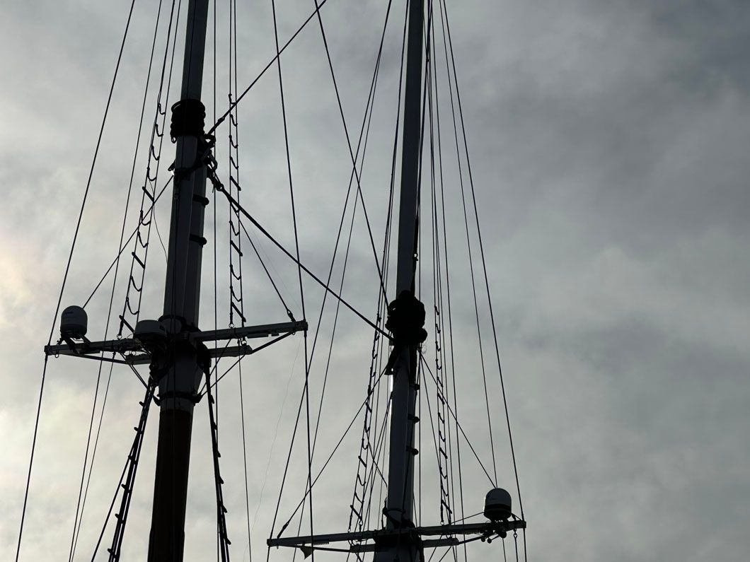 The masts of a sailboat are silhouetted against a cloudy sky