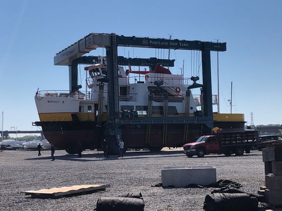A large ship is being lifted by a crane in a gravel area