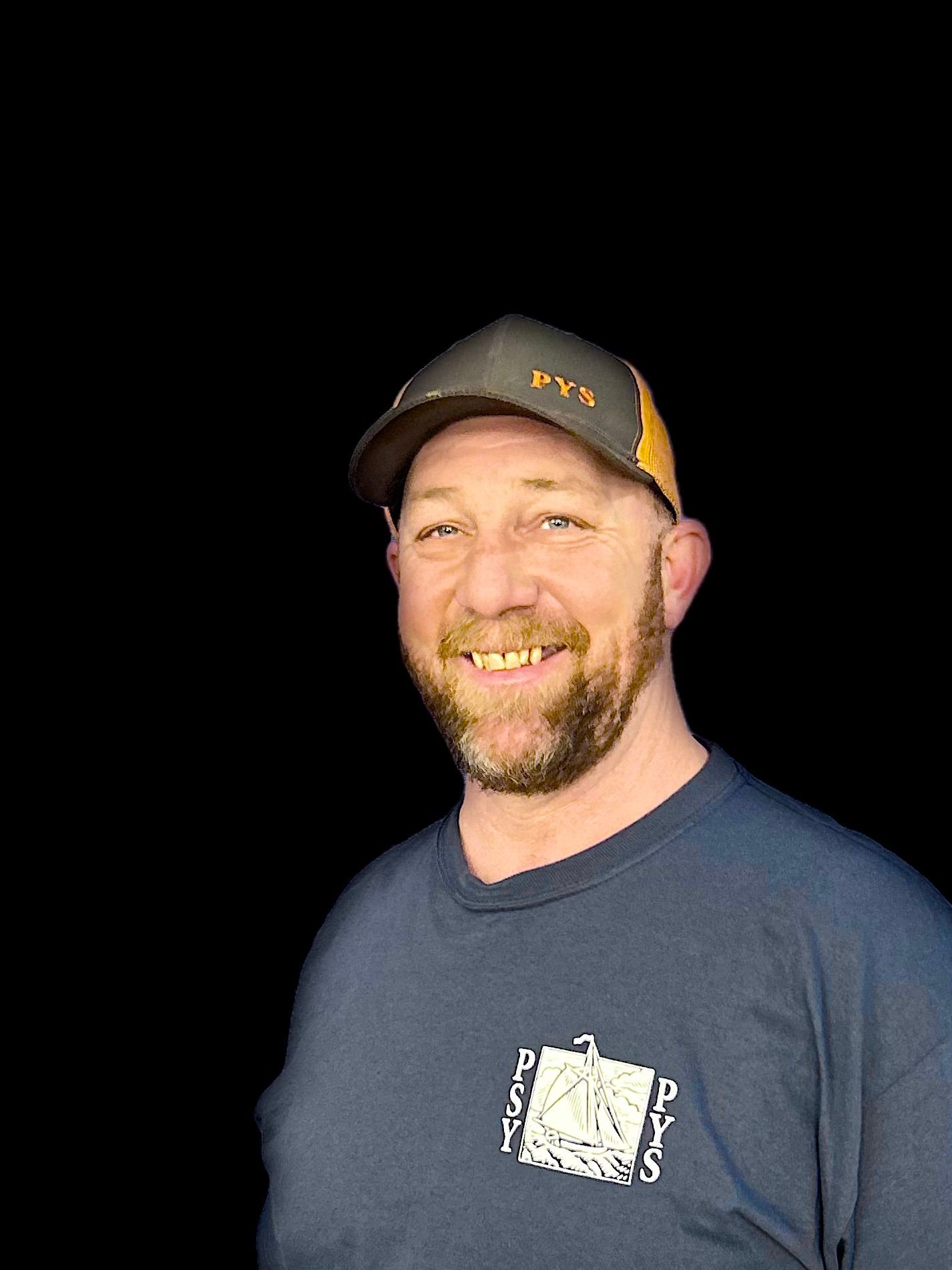 A man with a beard wearing a hat and a blue shirt is smiling.
