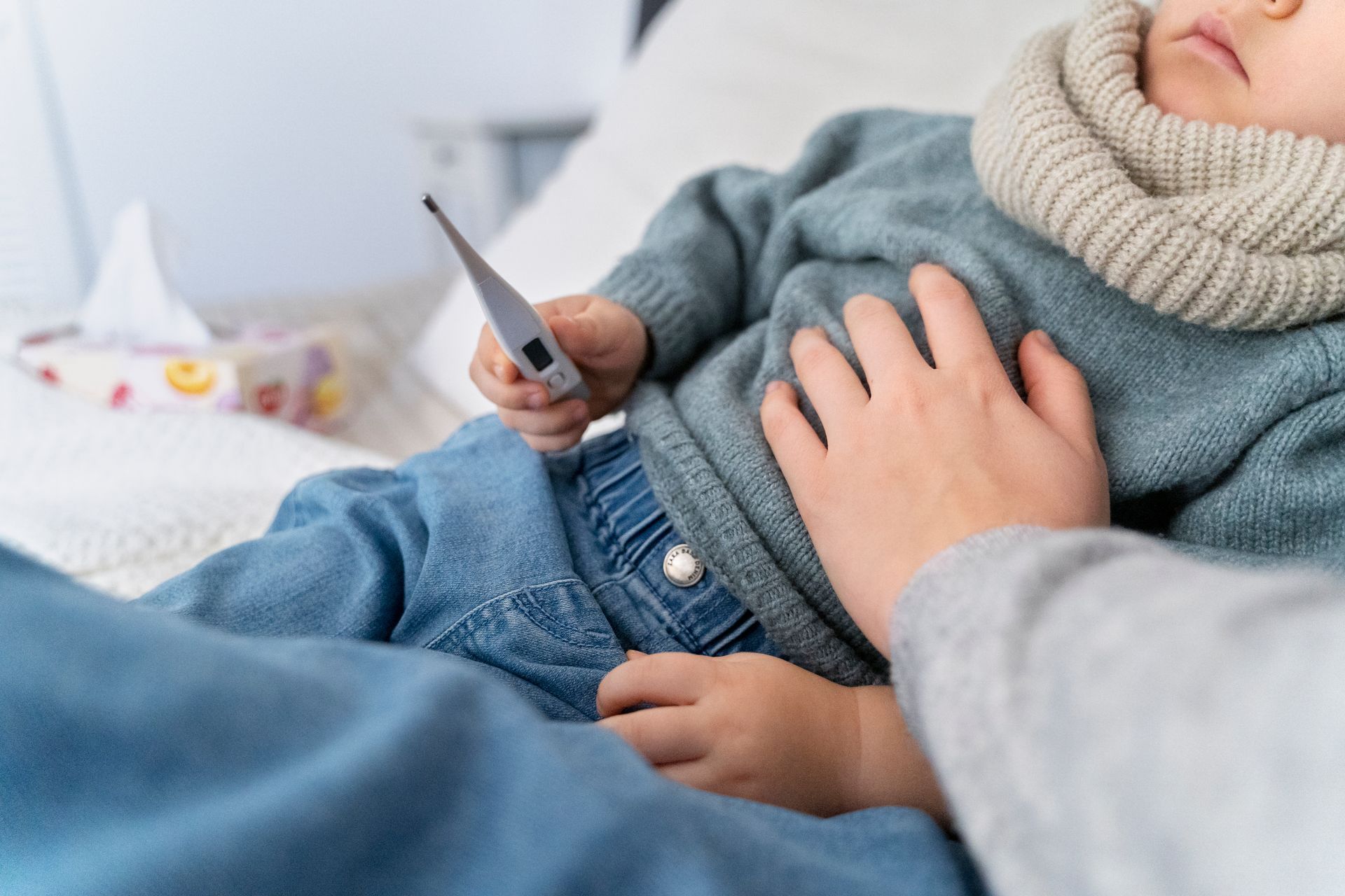 A toddler showing fever symptoms holds a thermometer while their caregiver monitors them.
