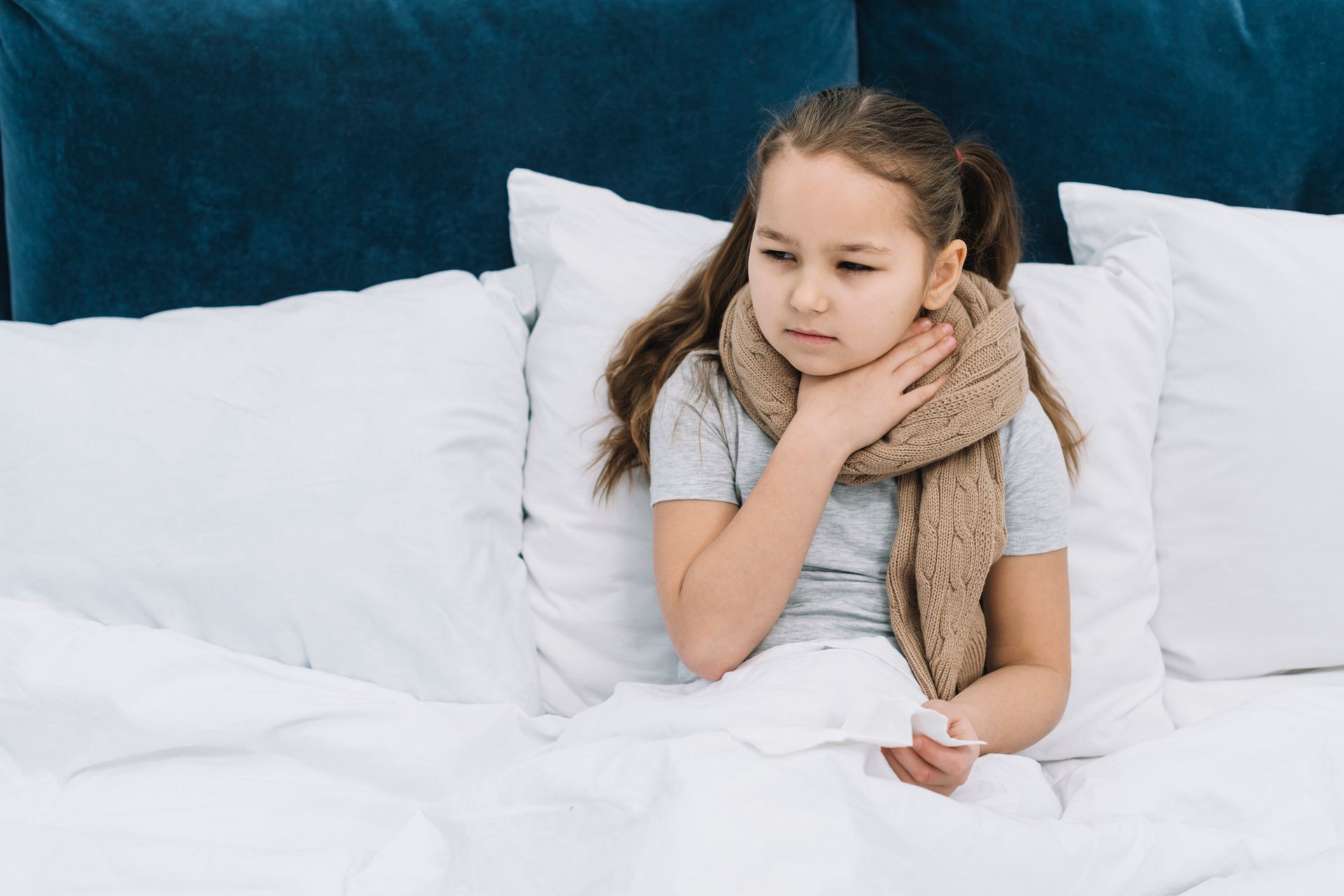 A girl sits in bed with a scarf around her neck,  indicating a sore throat.
