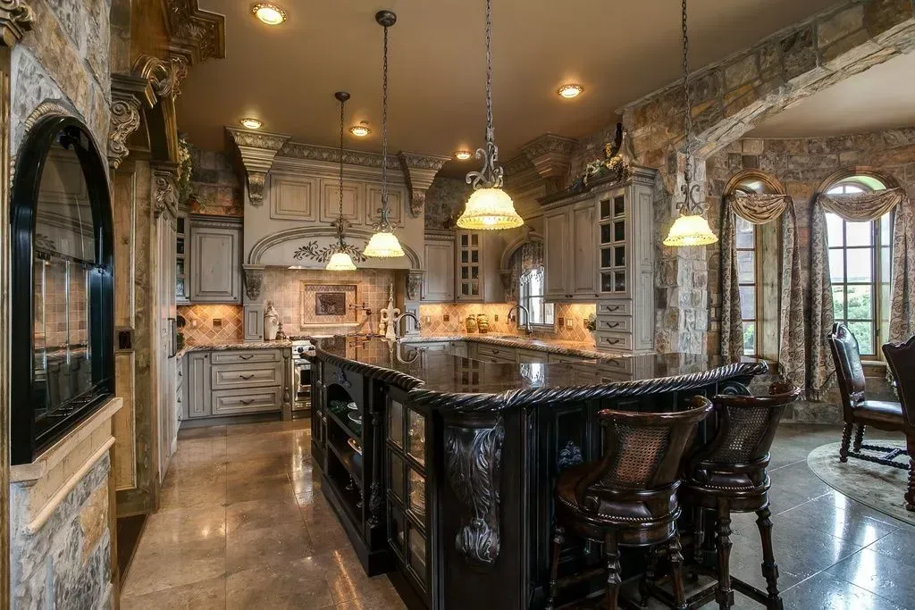 Ornate kitchen with dark island, light cabinets, stone walls, and pendant lights.