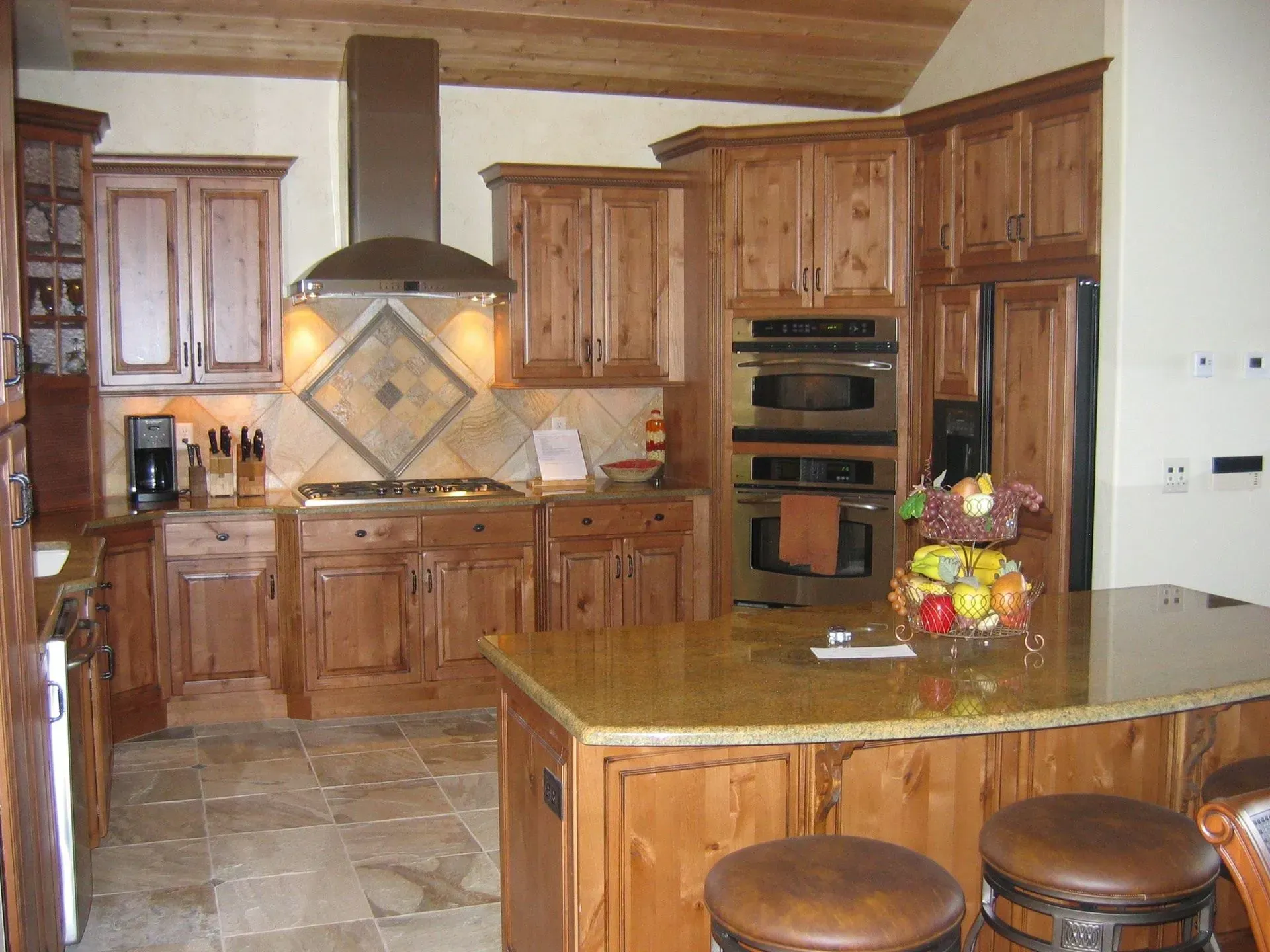 Wooden kitchen with granite countertops, island with stools, stainless steel appliances, and tile backsplash.
