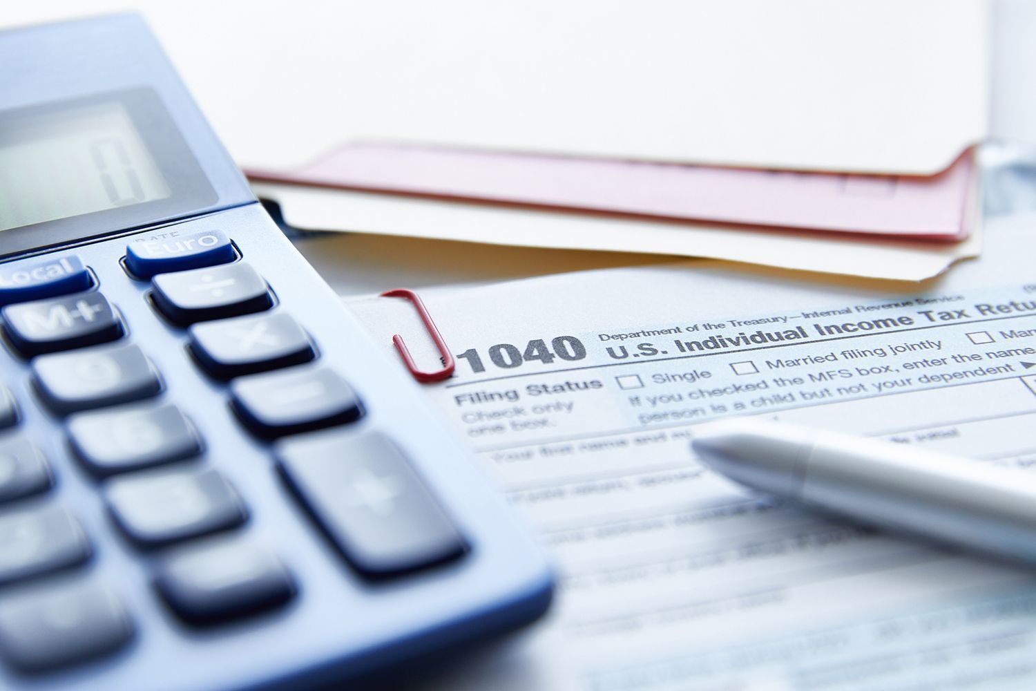 Calculator, pen, and tax forms with a red paper clip on a desk.