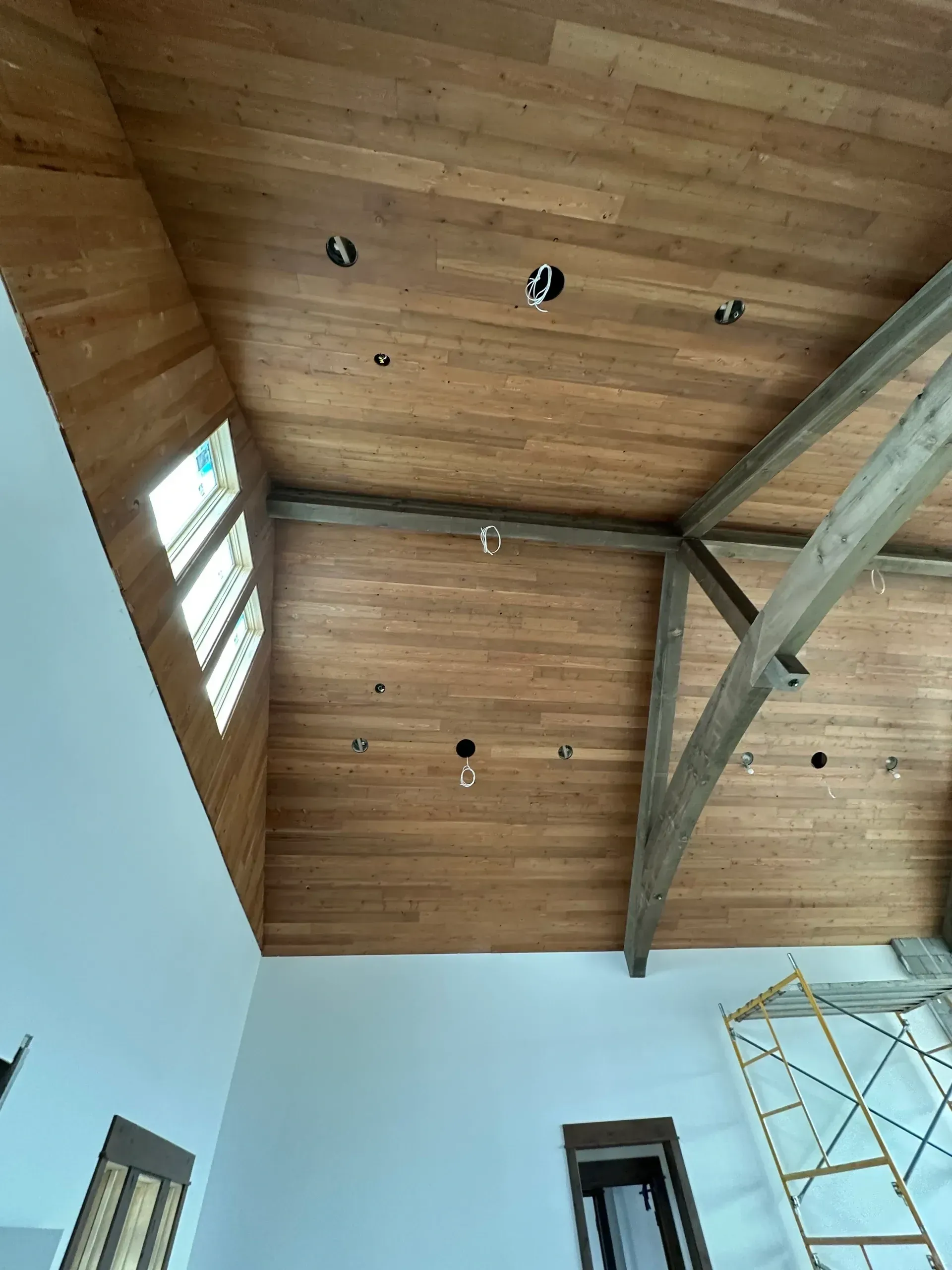 Wooden ceiling with recessed lights, windows, and exposed beams in a room with white walls and a glimpse of scaffolding.