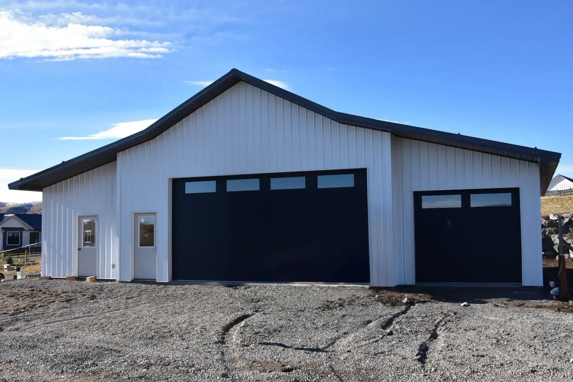 White barn-style garage with black doors and roof. Gravel driveway, blue sky.
