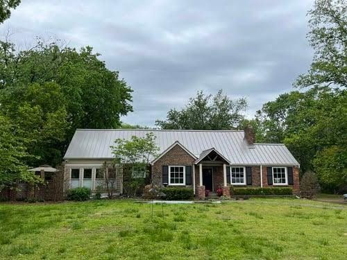 A brick house with a metal roof and black shutters is sitting on top of a lush green field.