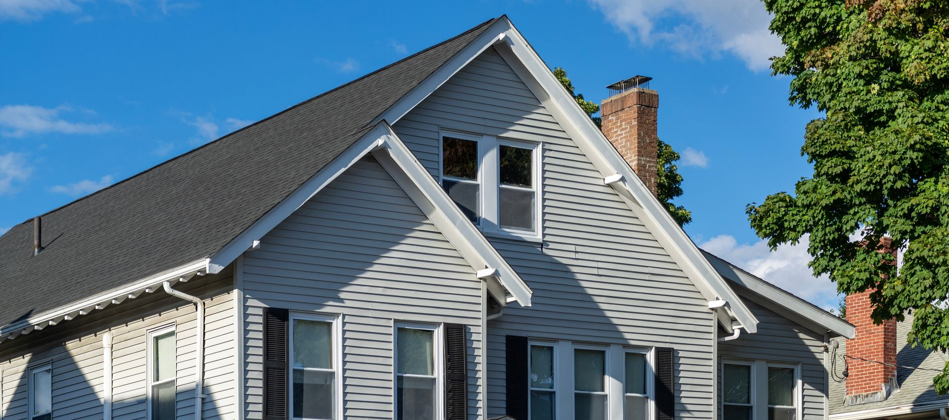 Two-story house with gray siding, black shutters, and dark roof against a blue sky with clouds.